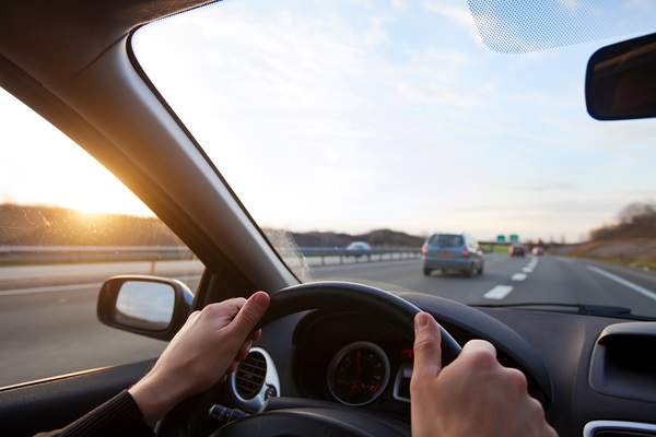 A driver's perspective from inside a car, holding the steering wheel while driving on a highway at sunset, with other vehicles visible on the road ahead.