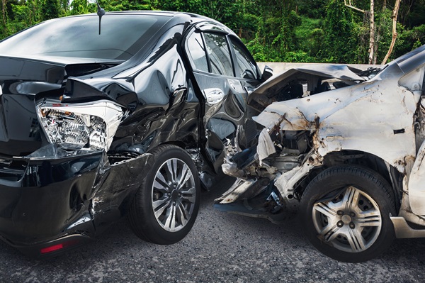 Two heavily damaged cars after a severe side-impact collision, with debris scattered on the road and a green wooded background.