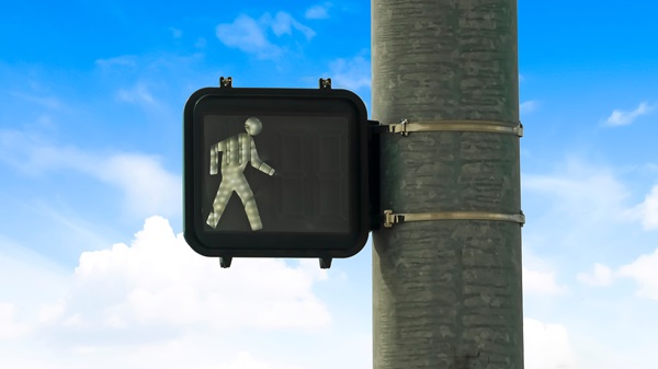 Pedestrian traffic light showing a white walking figure, mounted on a pole against a blue sky with clouds.