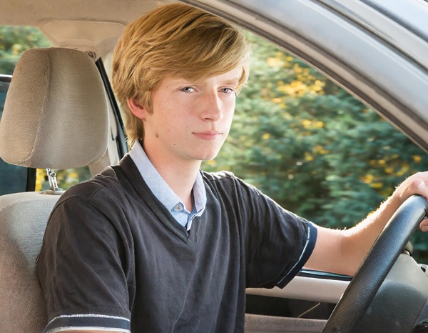 A teenage boy with blonde hair is sitting in the driver's seat of a car, looking directly at the camera with a serious expression. He is wearing a black shirt over a light blue collared shirt. His left hand is on the steering wheel, and he is sitting upright.